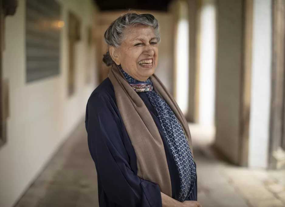 Smiling Pakistani woman with grey curly hair standing in a vaulted coridor.