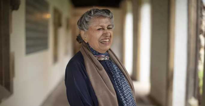 Smiling Pakistani woman with grey curly hair standing in a vaulted coridor.