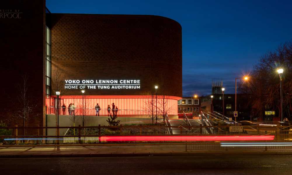 Outside a brick auditorium at night, there is traffic on the road. Illuminated text reads Yoko Ono Lennon Centre, Home of the Tung Auditorium.