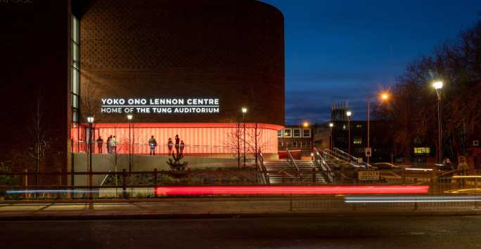 Outside a brick auditorium at night, there is traffic on the road. Illuminated text reads Yoko Ono Lennon Centre, Home of the Tung Auditorium.