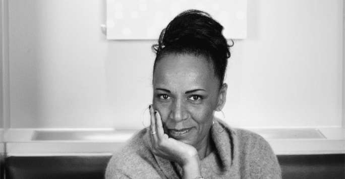 Woman with hair tied back leaning on a wooden table. Behind them is abstract painting.