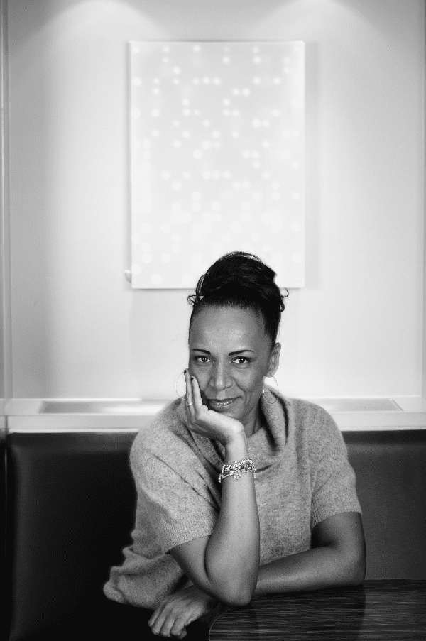 Woman with hair tied back leaning on a wooden table. Behind them is abstract painting.