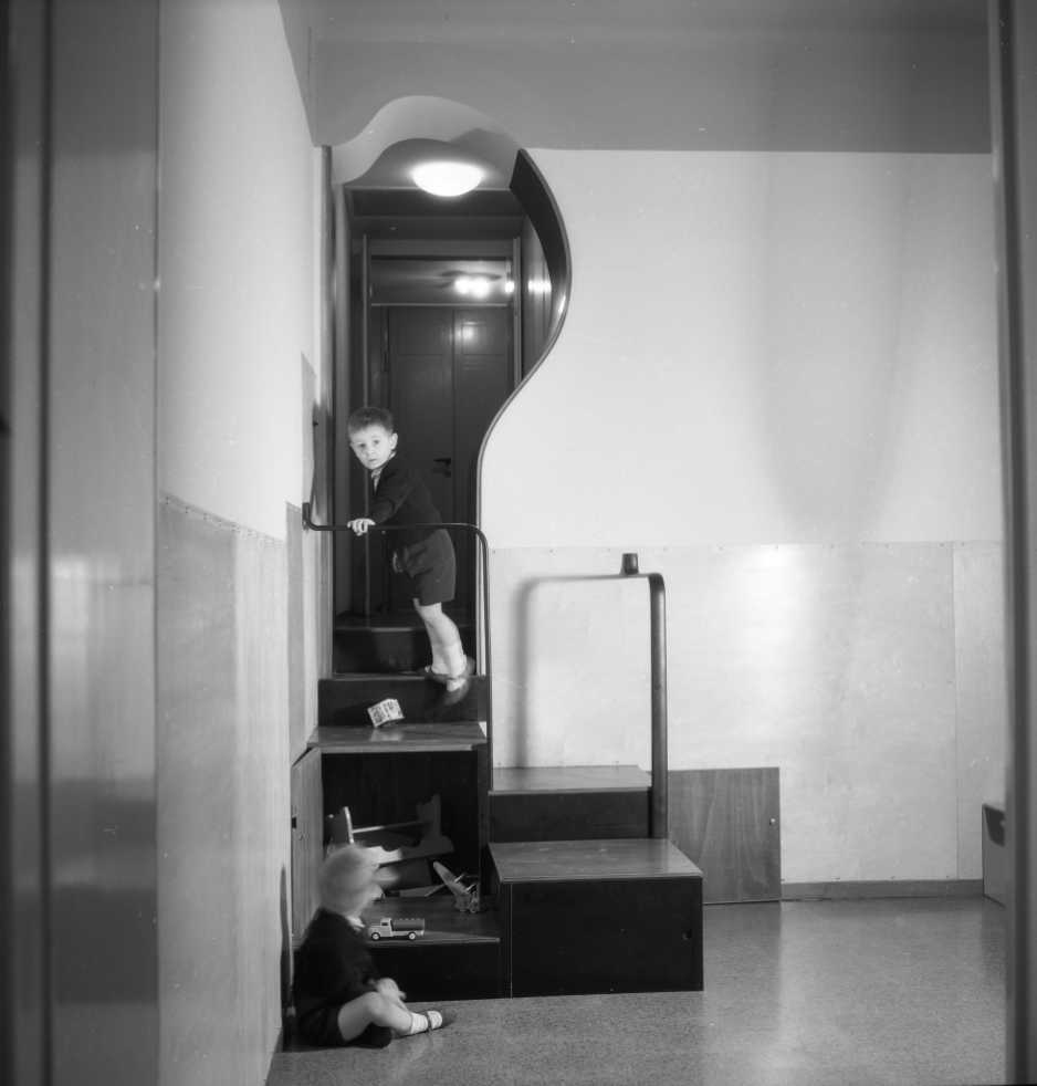 Male child wearing shorts standing halfway up a staircase, they have just dropped a cube with large letters on each face down the stairs.