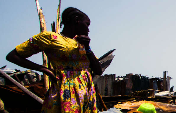 An African woman in a floral dress standing look at discarded kitchen bowl, behind her is housing built from corrugated iron.