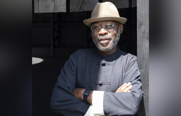 Smiling older black man with a grey beard, standing with arms crossed and leaning against the doorframe in a sunlit doorway. They are wearing a hat, jacket with upturned sleeves, and rimless glasses.