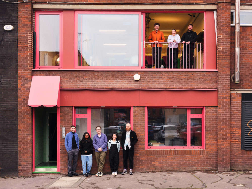Front of a brick building with red windows and doors. A mixed group of people are standing in front of the main door on the left hand side of the buidling.. Another group of people are standing in a large open window on the first floor.
