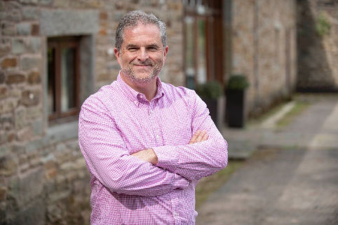 Middle aged man with short grey hair and a trimmed beard wearing a long sleave check shirt. They are standing outside, arms crossed, in front of a stone built domestic building.