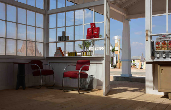 Stylised photo of the interior of a garage office in the American mid west. Two chrome and leather chairs sit in front of a wooden walls with a wood framed window, an open door leading out to a dusty road. On the otehr side of the road a diner can be seen, in the distance a mountain. The sky is blue with little fluffy clouds.