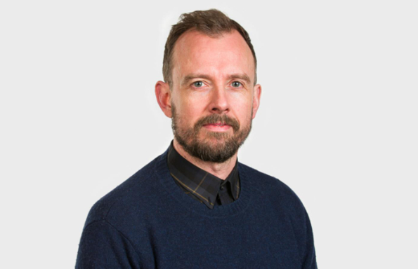 Middle aged man with short cropped hair and beard, wearing a dark tartan buttoned up shirt and jumper.