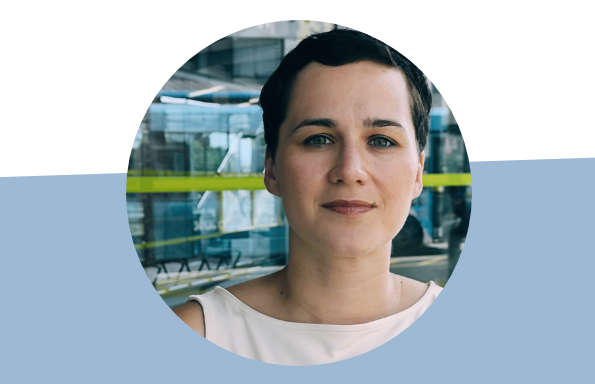 Circular graphic element containing a photograph of the head and shoulders of a woman with short hair and wearing a white t-shirt, they are standing in front of a glass fronted building.