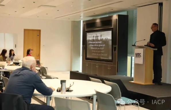 A middle aged man standing at a lectern speaking to a conference room. Two his right is a display screen showing slides, delegates site round tables watching him.