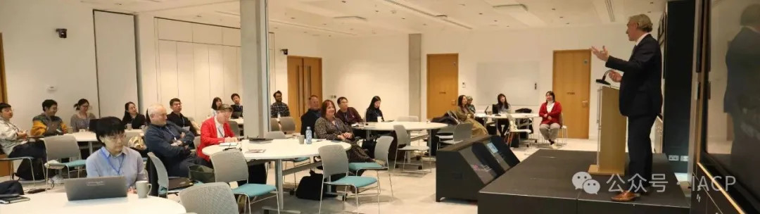 A middle aged man standing at a lectern speaking to a conference room. Two his right is a display screen showing slides, delegates site round tables watching him.