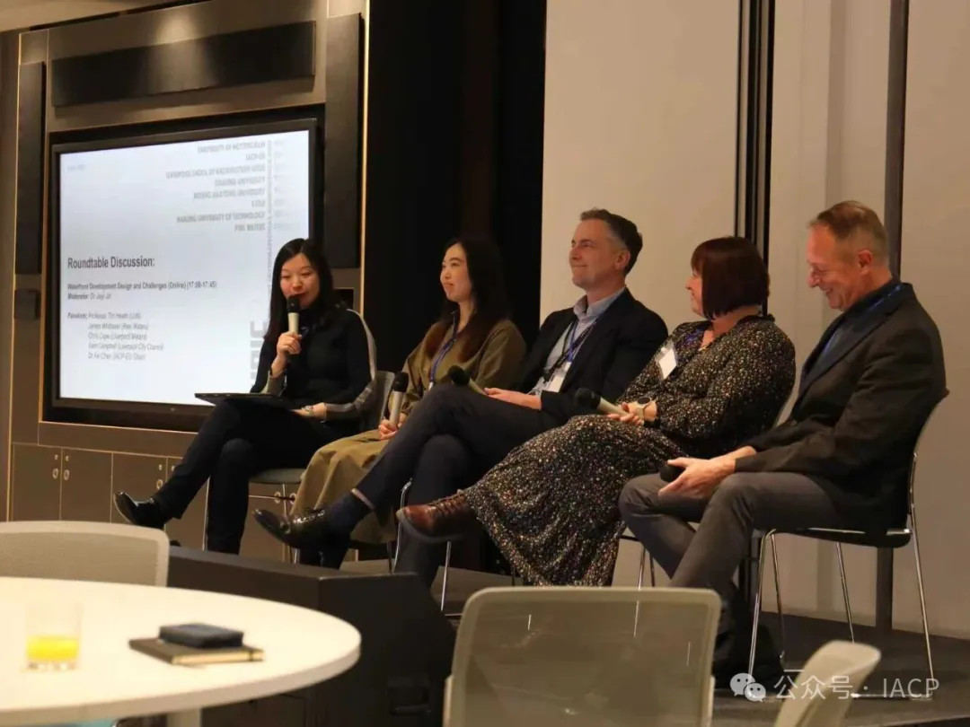 In a conference room three women and two men are sitting on a stage speaking to an audience. One of the women is holding a microphone and reading from a tablet computer.