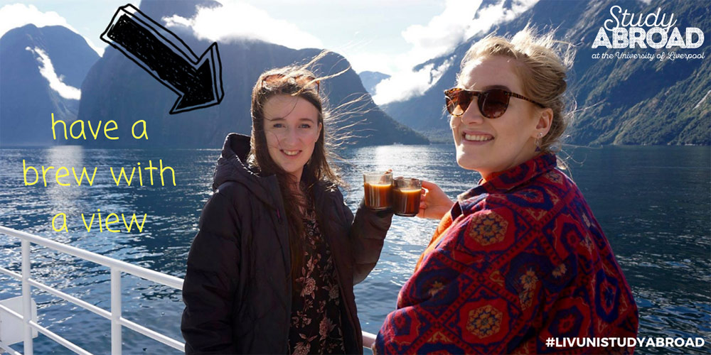 Two young women leaining on railings of a ship, sailing through a fjord. Both are dressed for cold weather and holding cups of tea. Mountains can be seen in the backgroung. Overlayed is text reading 