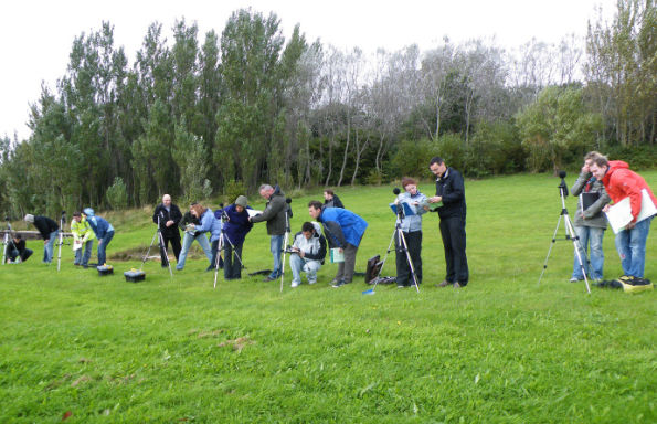 A group of people standing on a grassy hillside with sound measuring equipment mounted on tripods. A lecturer is walking around instructing them. Behind them is a row of short trees and bushes.