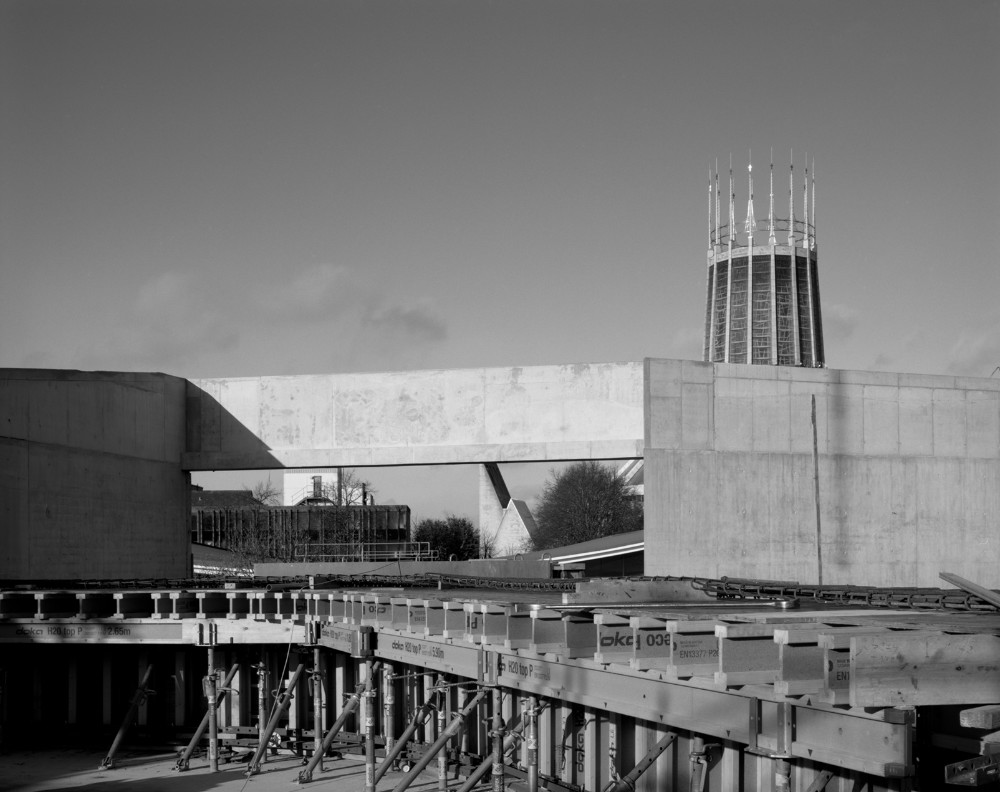 Black and white photo of a half completed building. In the foreground is concrete shuttering, in the distance the spire of a modern cathedral.