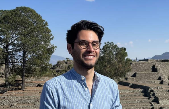 An hispanic man with short dark hair, beard and glasses wearing a blue collarless shirt is smiling and standing in front of a South American pyramid