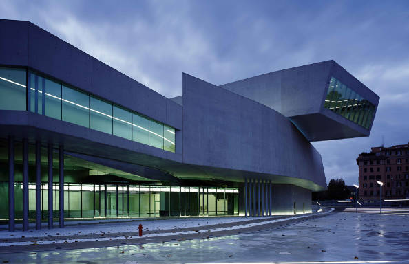 Modernist building in twilight, the concrete walls are sightly curved and with illuminated windows along part of their length. A second floor sits at an angle above the main body of the building.