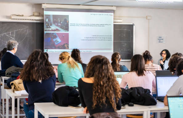 A woman with shoulder length hair and glasses is sat at a desk in front of a class of students lecturing. On the screan behind her is a slide that reads 