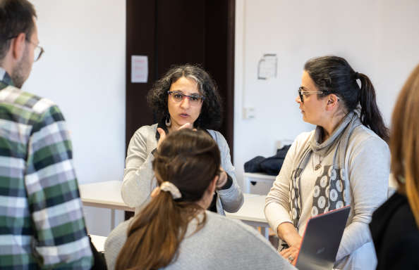 A group of people are discussing a lecture, a woman with shoulder length hair and glasses is gesticulating with her hands.