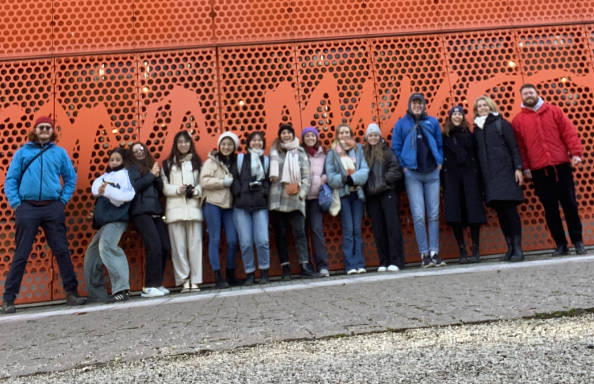 A group of students and staff standing on a concrete pavement, in front of a perforated orange metal wall. All are dressed for cold weather. The photograph is taken at an angle.