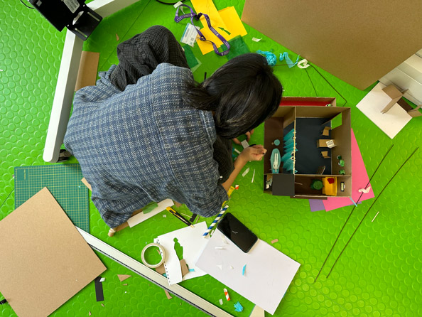 View looking down on a student making a model, they are surrounded by card, paper and tools