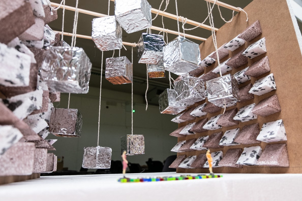 Model of architectural space. The walls are covered in angled panels made from foil, foil cubes hange from the ceiling. Two small mod