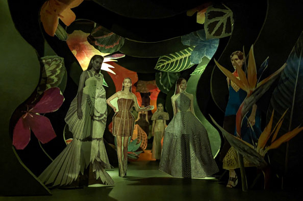 Cardboard cutouts of white fashion models set against a miniature theatre set with 2D flowers