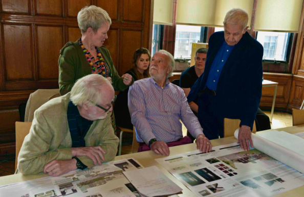 In a wood panelled library two bearded older white men sit at a table, one is examining an architectural drawing, the other is chatting to a grey haired woman standing behind them. To their right a third white man looks at the drawing. Behind them are a female and a male student.