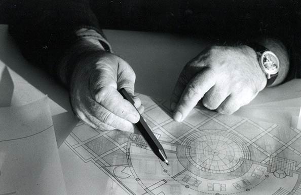 Two hands, one holding a mechanical pencil, resting on a plan of a building