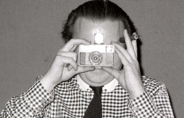 Middle aged man in a check shirt holding up a Kodak Instamatic camera and a cigar. The flash bulb is going firing. Credits: Cedric Price with an Instamatic camera 1972 (Kathy de Witt/RIBA Collections)