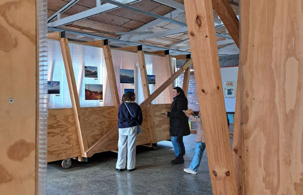 Two women looking at an exhibition of photographs. The Photographs show city scenes from around the global south and are mounted on a plywood structure.