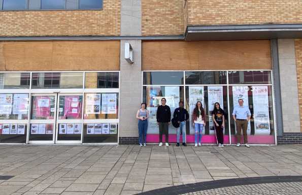Students standing in front of the glass doors of a modern brick building. There are posters on the windows.
