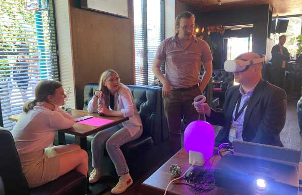 Inside a restaurant two women are sitting at a table watching a man in a suit wearing VR goggles.