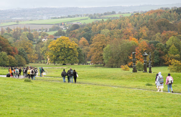Grass covered hillside with a pass leading down to tree lined river at the bottom. Students are walking away from the camera.