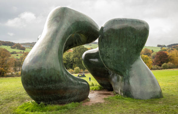 Large bronze henry Moore scuplture in two parts, sitting in farmland. between the two parts another sculpture can be see in the distance.