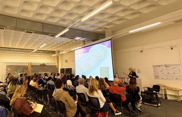 A woman addressing a group of seated people in a lecture theatre. A presentation on architecture is projected onto a screen in front of them.