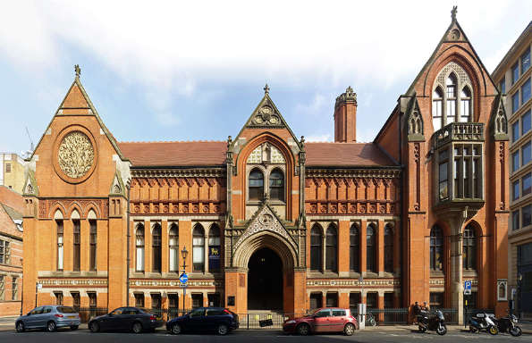 Front of a red brick Victorian Gothic building with cars lined up on the street in front.