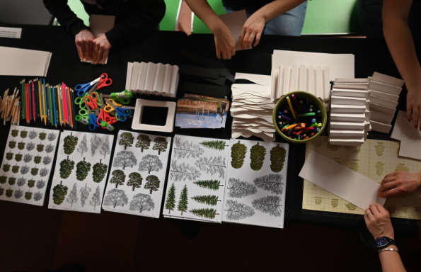 Overhead view of a table with neatly arranged craft supplies including paper, scissors, pencils and pens, with photographs of trees and landscapes ready to be cut out. Several sets of hands can be seen working with the materials.