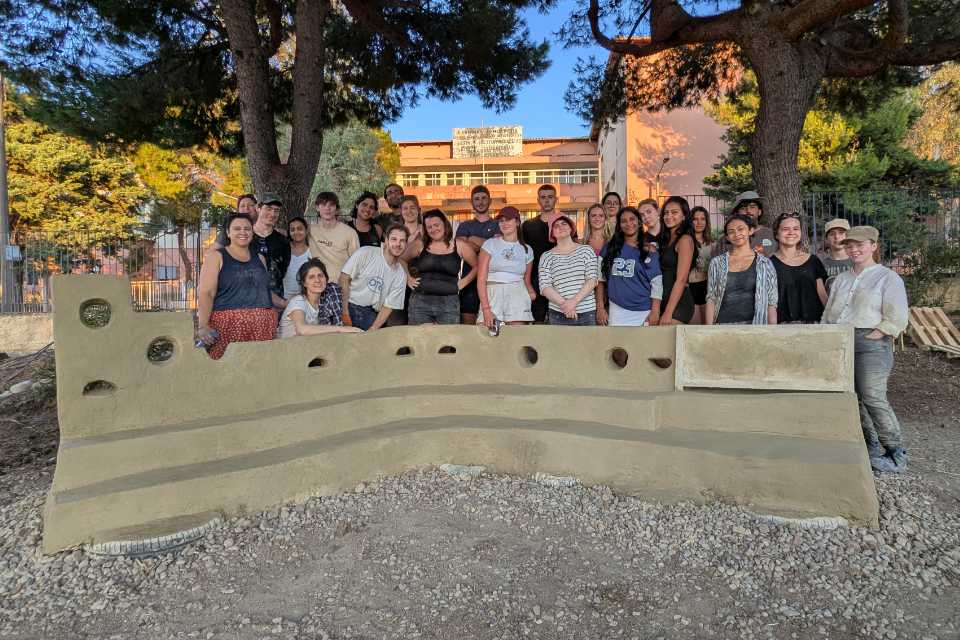 Group of people standing beneath a pair of trees in the sun. Behind them is a rectilinear two floor school building. In front of them is a curved wall with built in seating.