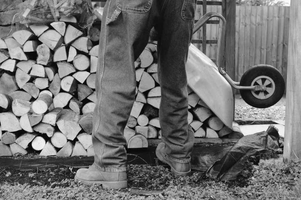 A pair of trousered legs standing in front of a chopped wood pile. A wheelbarrow rests against the logs.