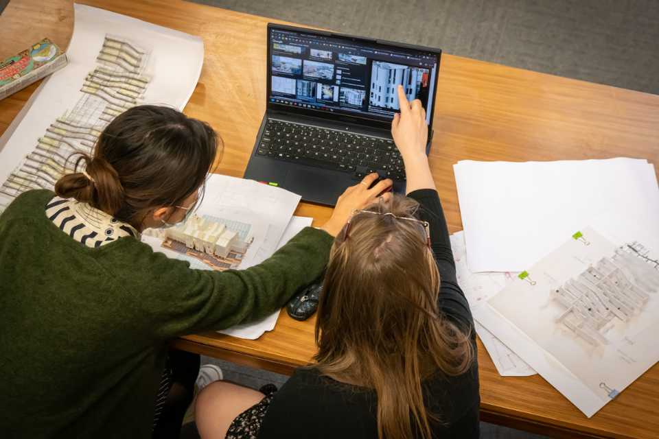 Two women sitting at a desk with an open laptop in front of them. One is pointing at an image of a housing block on the screen whilst the other uses the keyboard. One the table are plans and models of housing schemes.