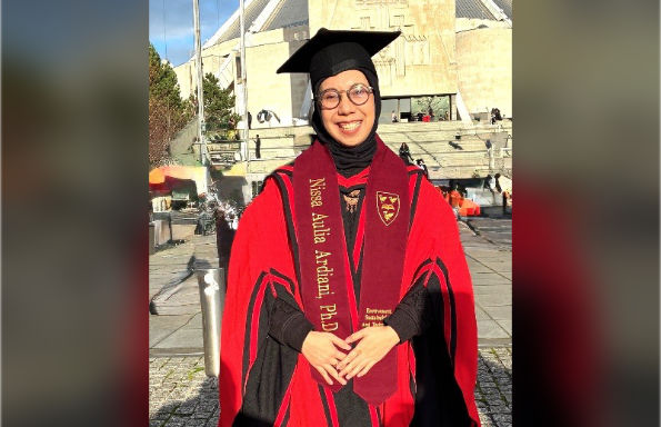 Woman in red and black academic gowns and headscarf smiling and standing in front of a modernist cathedral in the sunlight.