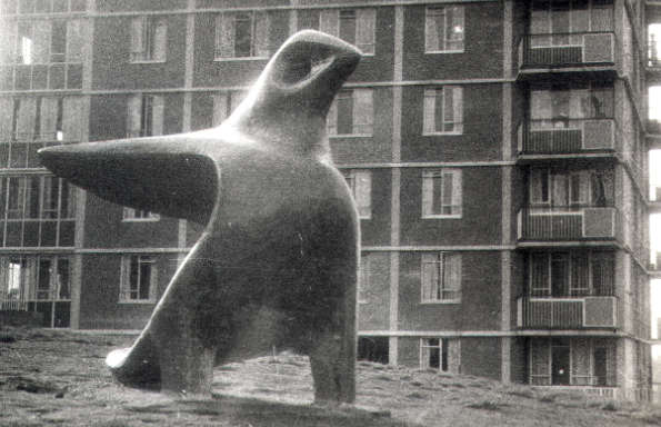 Vintage black and white photograph of a large abstract sculpture of a bird set in a grassed area outside a brutalist tower block.