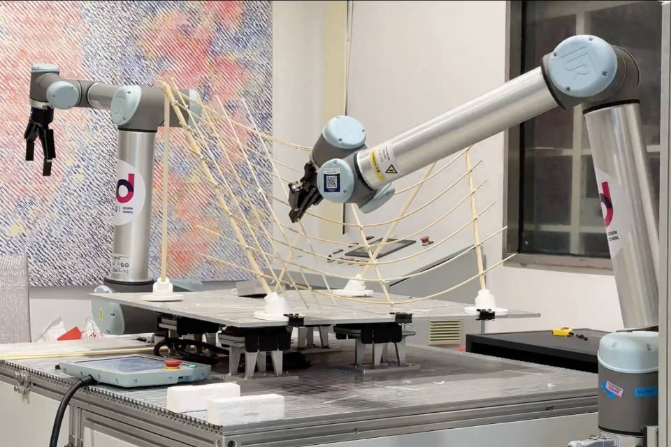 Two robot arms in a small room with a bamboo roof structure behind them. One is moving a bamboo rod into position on the structure.