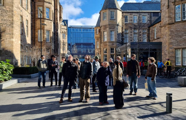 Group of men and women standing and looking up in a courtyard surrounded by imposing stone gothic buildings.