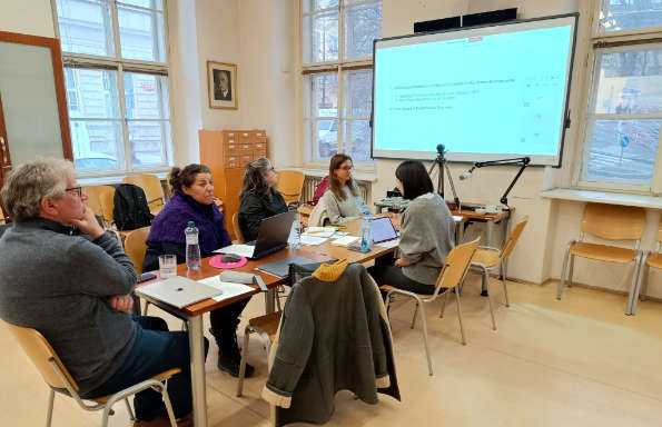 Men and women sitting around a table and looking at a projected screen. The room has a high ceiling and stone built buildings can be seen outside through the windows.