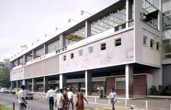Three-quarter view of a large modernist retail building in Africa. In front is a busy street with cars passing in front and pedestrians.