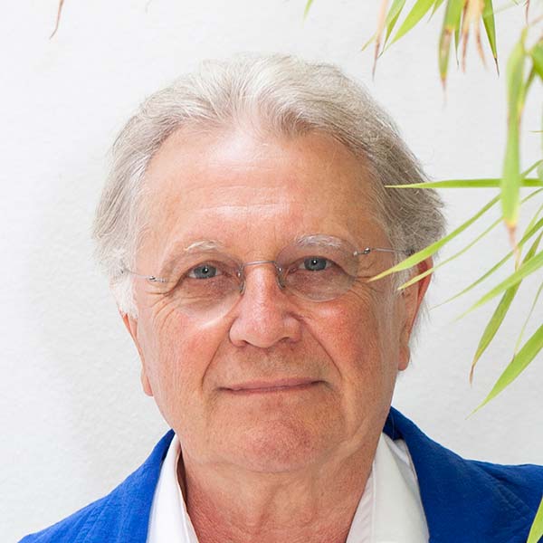 Middle aged man with white hair wearing a blue suit and white shirt standing in front foliage