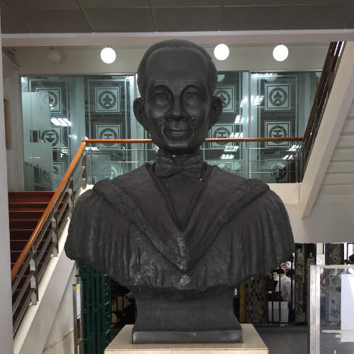 A dark cooper bust of a man wearing a bow tie and a gown, on a marble pedestal. The bust is placed in front of a grand entrance stairway.